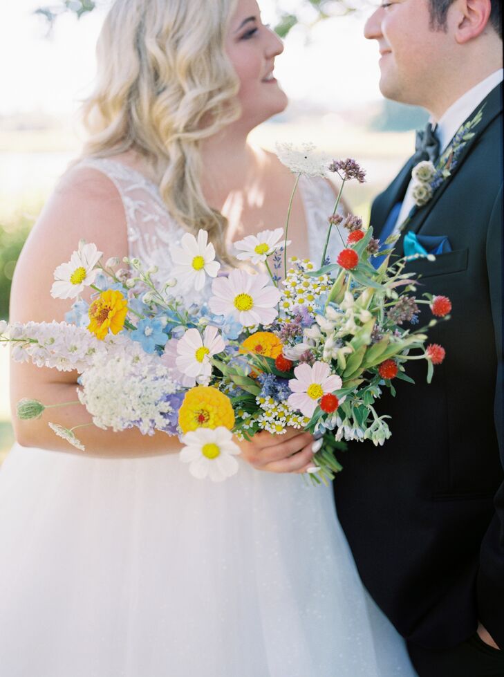 A bride in a tulle wedding dress and a groom in a black-and-blue tuxedo hold a bouquet of bright, colorful wildflowers in shades of yellow, blue, red, pink and white.