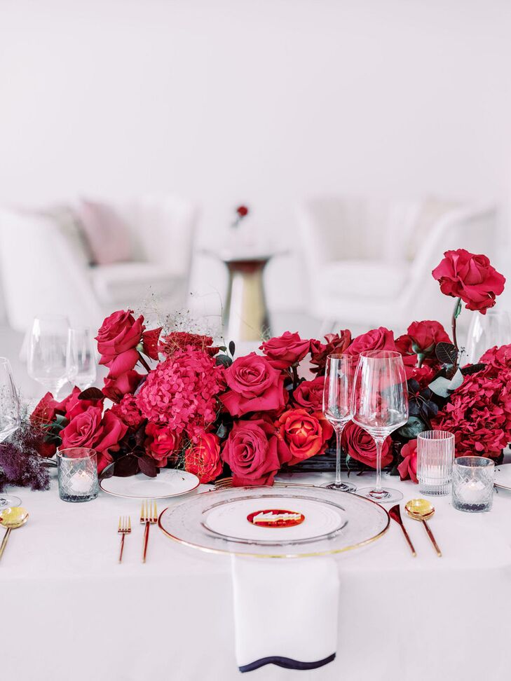 An all-white table setting with pops of red color in the floral table runner, along with gold details in the flatware and custom place card for a modern and elegant wedding rehearsal dinner.