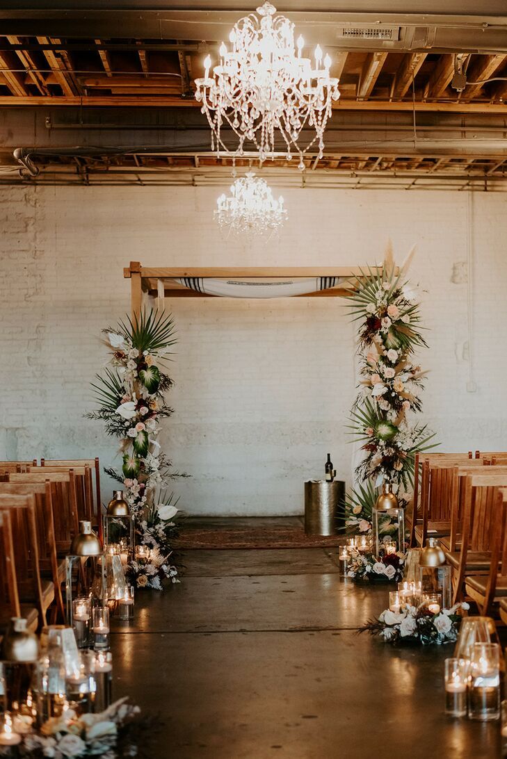 Palm-covered chuppah in industrial loft under ornate chandeliers. 
