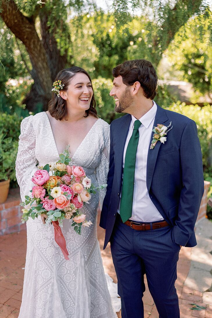 A bride in a lacy sheath gown with a plunging V-neckline and long sleeves with a fresh flower hair clip and a pastel bouquet smiles with her groom in a deep blue suit and a forest green tie with a coordinating boutonniere before their Southern California wedding.