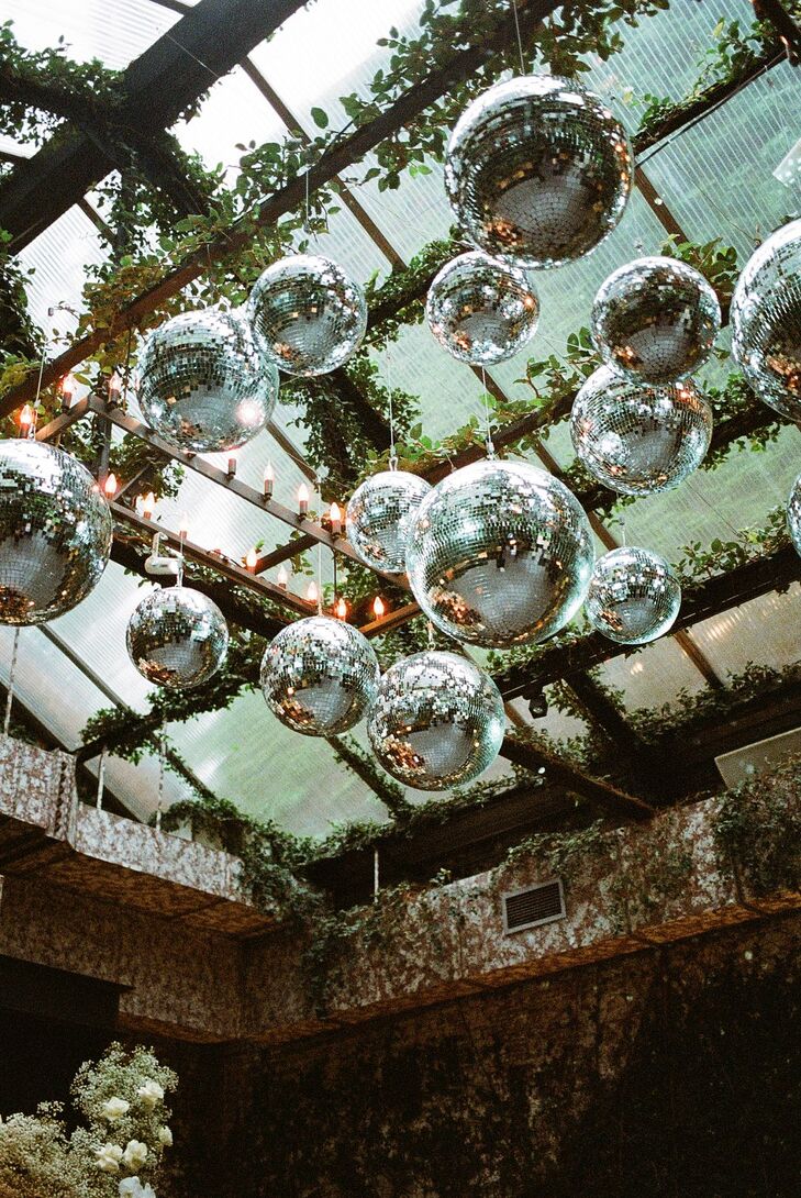 A custom disco ball chandelier installment flanked by roaming ivy/greenery and additional lighting suspended under a greenhouse-like skylight ceiling and overhead of the reception's dance floor at a modern wedding in Long Island City, New York.