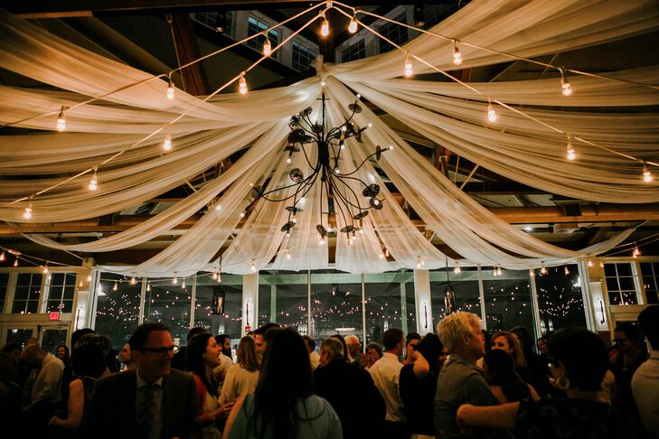 Originally, Janel and Charlie wanted to hold their reception in a more open environment. But with his family visiting from Seattle, Washington, it would be too much travel for their guests. So they opted for a loose lighting design. Building on the floor-to-ceiling windows and fabric along the ceiling at Liberty House Restaurant in Jersey City, New Jersey, they highlighted the space with delicate string lighting across the modern chandelier. Every detail made the room seem much larger and natural, as if they were partying below the stars.