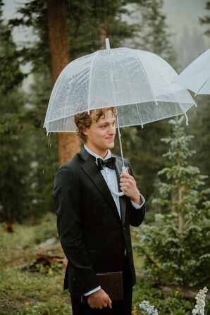 Groom in Classic Black Tuxedo Standing Under Clear Umbrella With Vow Book at Outdoor Ceremony