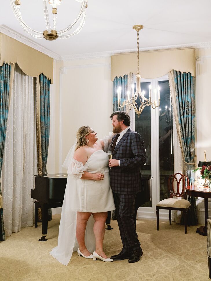 A bride in a white minidress, detachable sheer puffed sleeves, a white bridal cape and a small veil with drop earrings and small heels embracing her groom in a plaid, blue custom-made suit in a hotel suite before their elopement.