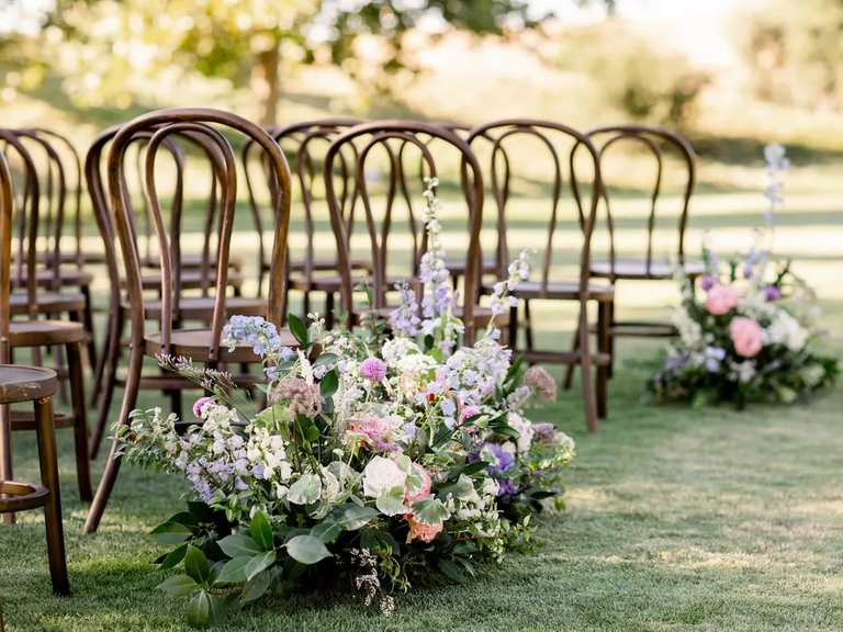 A springtime floral arrangement in pastel colors sits beside the wooden chairs of a wedding aisle.