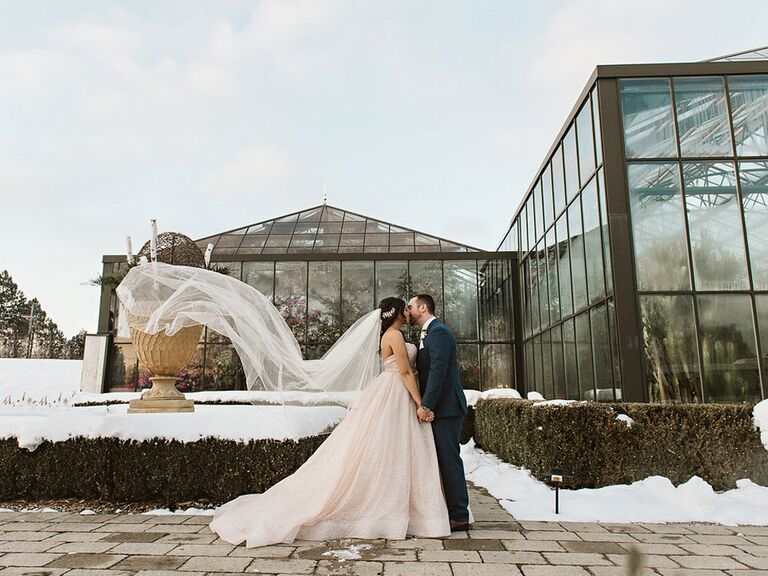 Bride and groom kissing on wedding day at outdoor venue with veil blowing in the wind