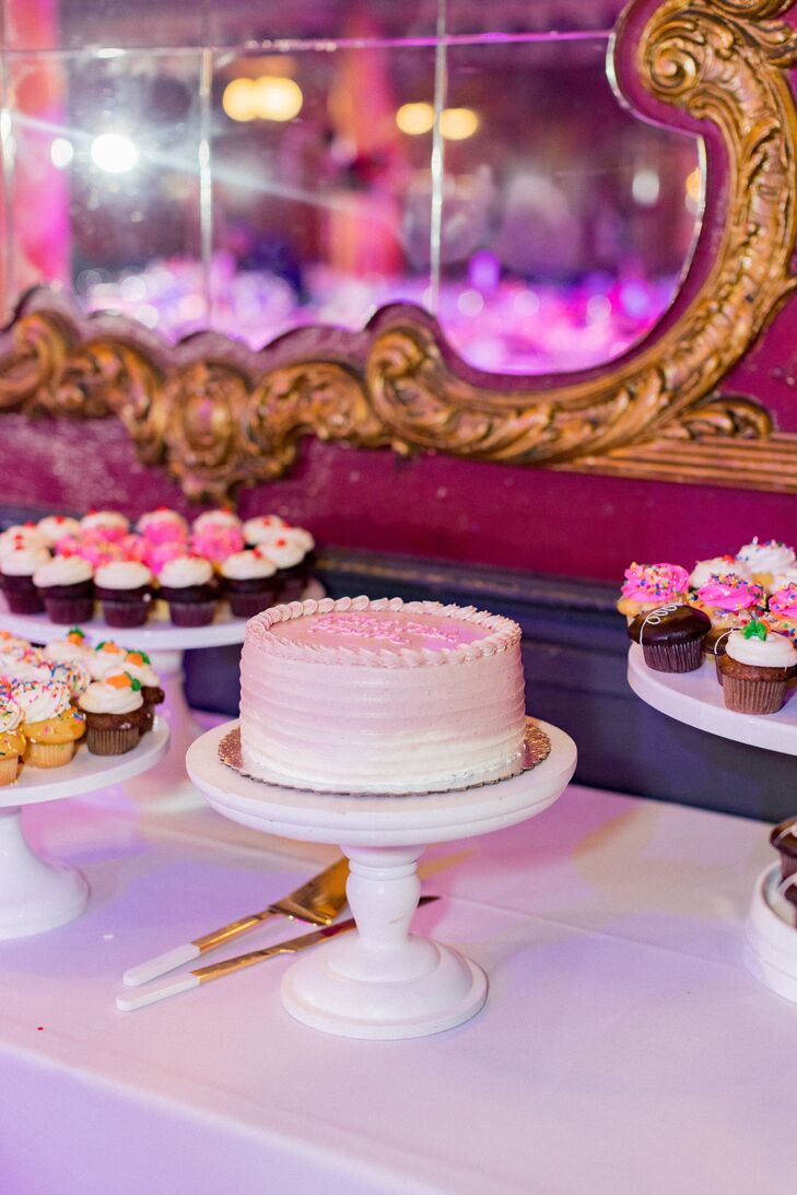 A single-tier wedding cake in an ombre white-and-pink palette on a dessert table filled with tiny cupcakes in pink and white.