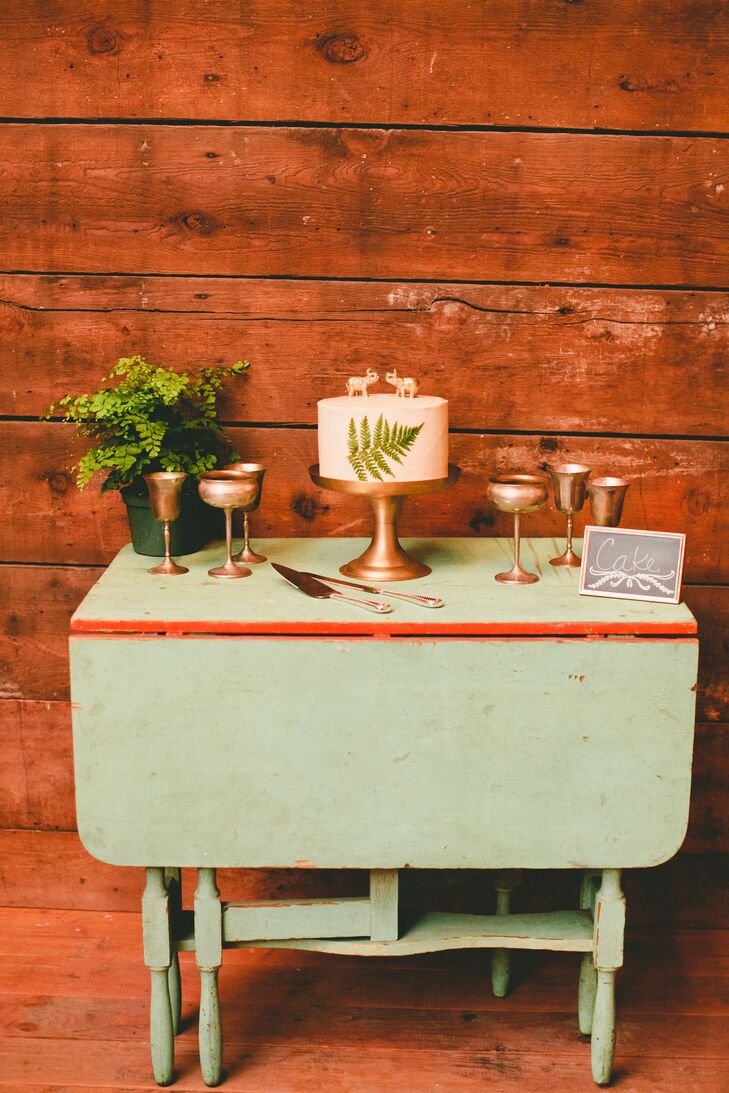 The single-tier white wedding cake with a fern-leaf design on the front had a topper depicting two gold elephants. The cake sat on top of a gold cake stand, surrounded by other gold pieces decorating the green display table.
