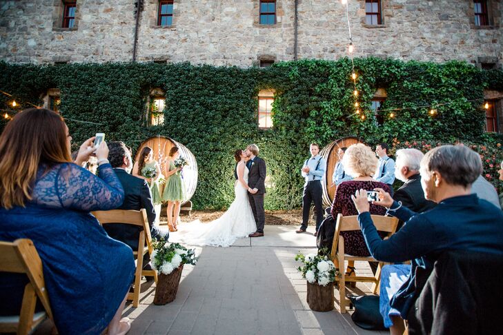 Michelle and Ryan had their ceremony outside at the Culinary Institute of America at Greystone in St. Helena, California. They walked down the aisle lined with arrangements of ivory blooms and greenery positioned on top of logs. The first kiss was shared in front of an ivy-covered wall backdrop, surrounded by twinkling string lights.