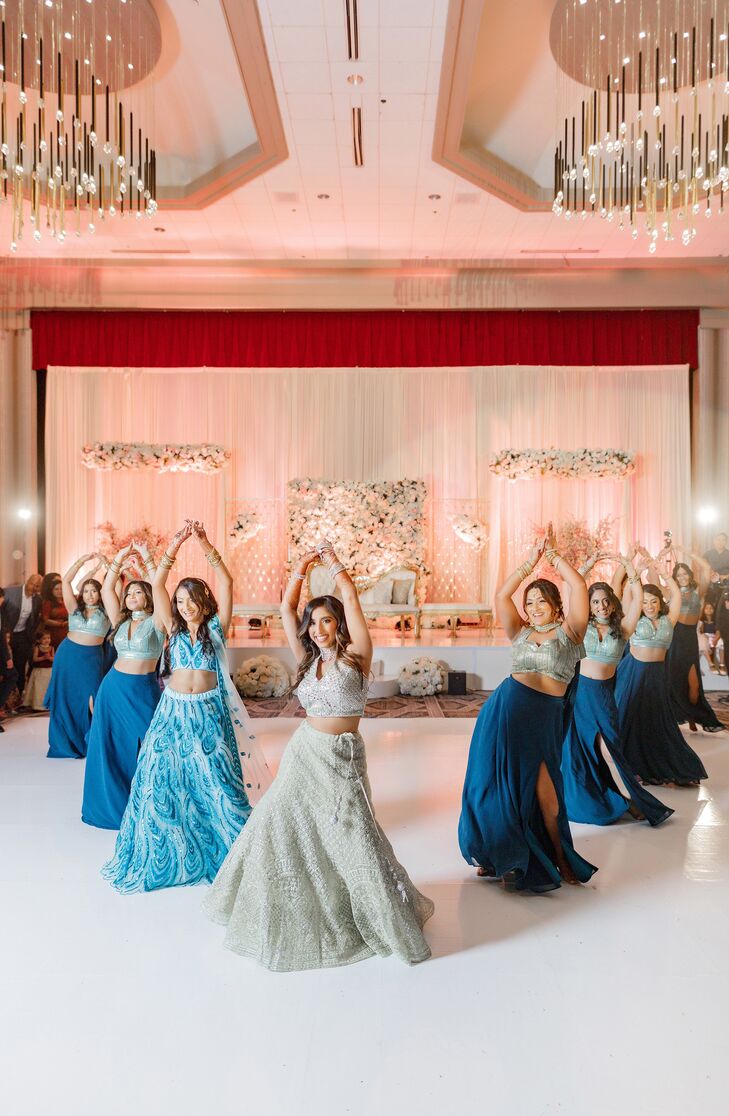 A bride in a beautiful, hand-embroidered white bridal lehenga and a down hairstyle performs a choreographed dance with her bridesmaids (in blue lehengas) on the dance floor at her elegant, glamorous Knanaya Indian Christian wedding reception.