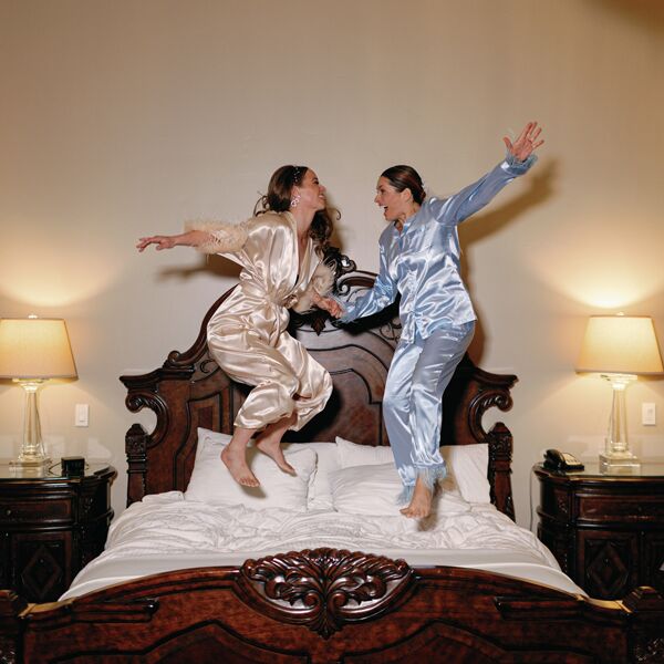 Two people in silky pajamas jumping on an ornate wooden bed.