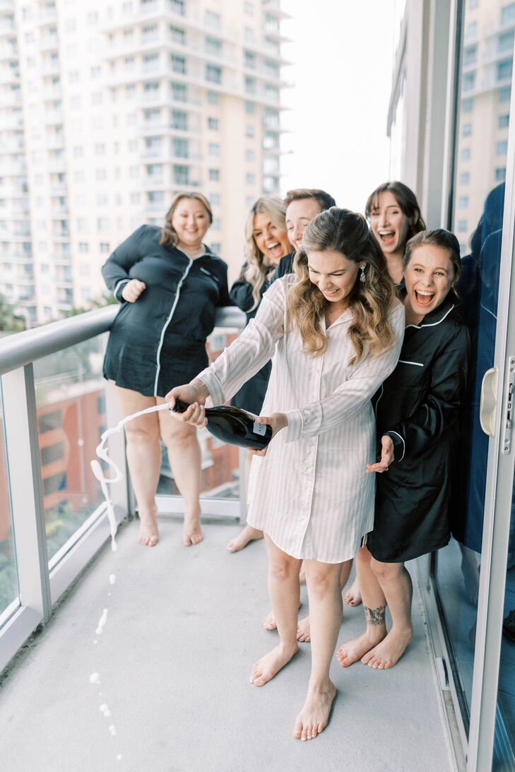 A fun bride in a long, white, getting-ready dress shirt popping a bottle of Champagne for her bridesmaids/wedding party in silk, black dress shirt on a high-rise balcony in Miami, Florida.