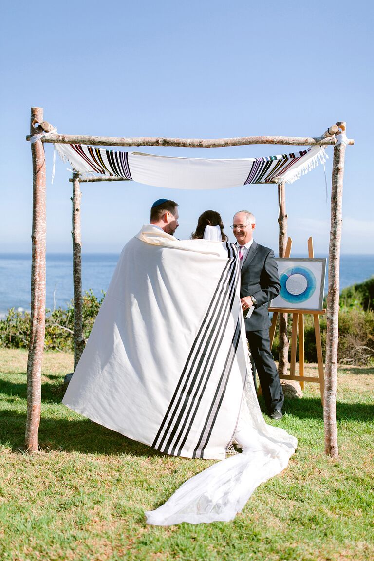 Couple wrapped in prayer shawl underneath birch chuppah.