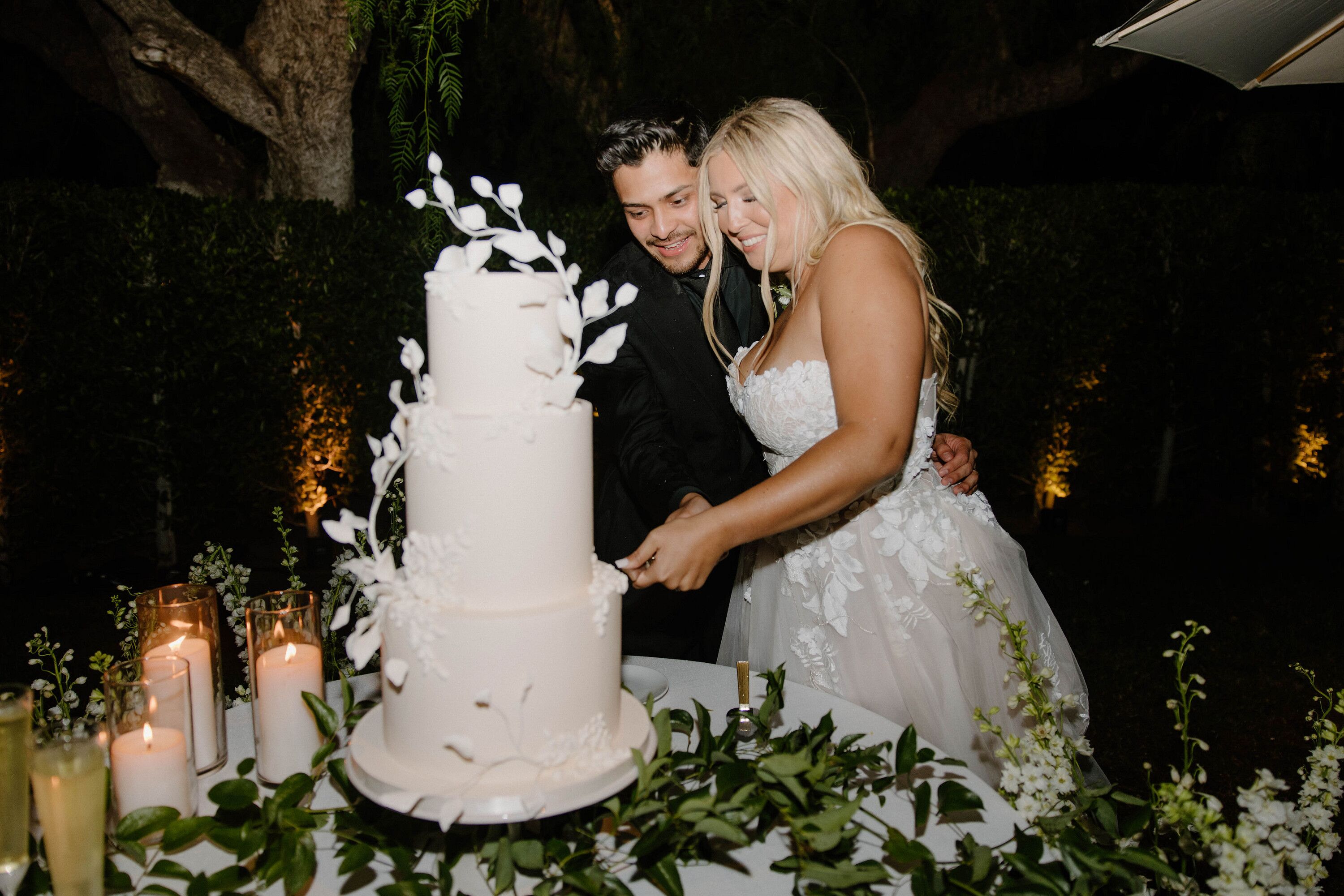 Bride and Groom Cutting Three-Tier Cake With Floral Appliqués, Greenery and Candles