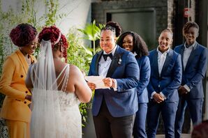 Bride in Blue Suit Reading Vows to Her Bride With Wedding Party Behind Her