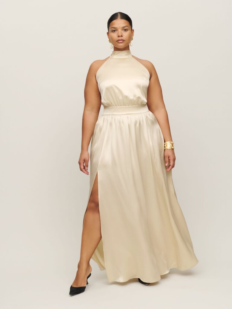 Model wears a cream colored dress with a high neckline and belted waist. 