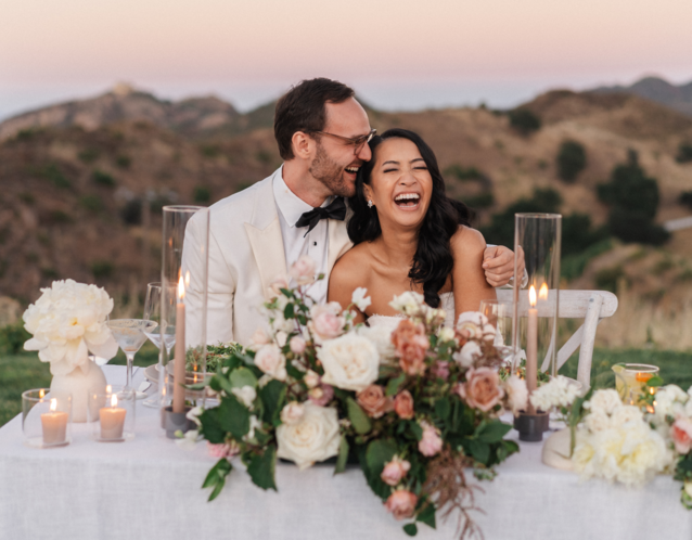 Couple sitting at reception table laughing with mountain background
