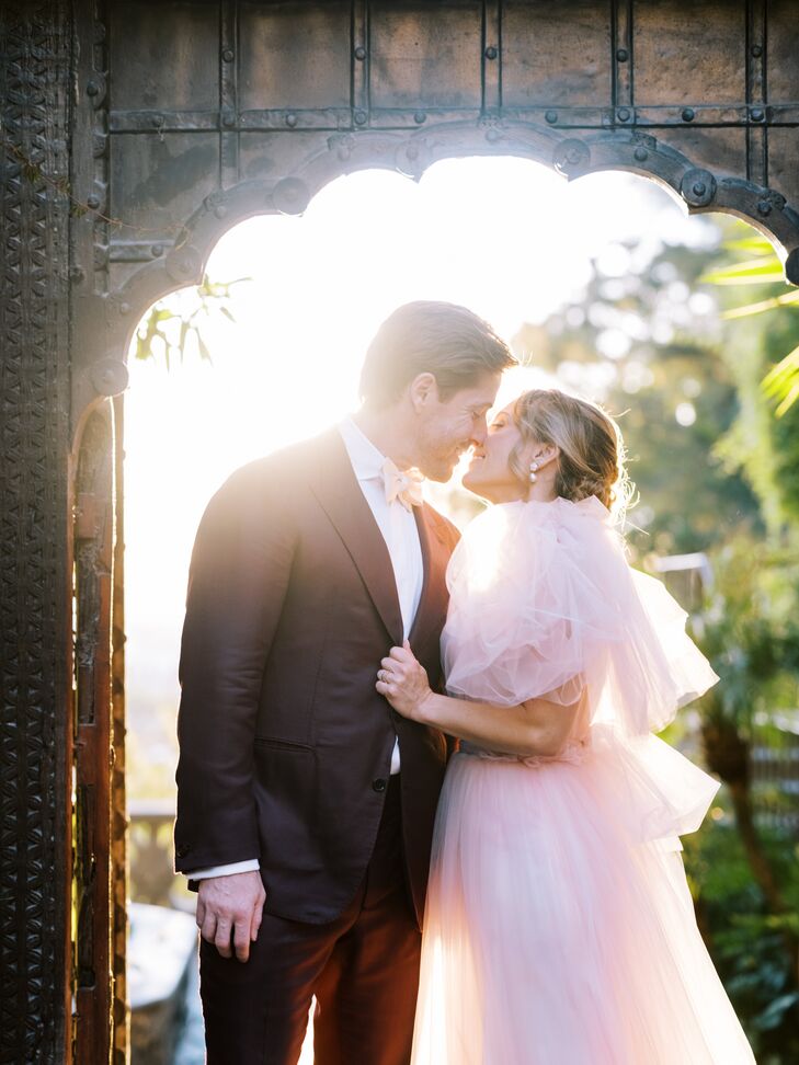 Groom in Maroon Suit Kisses Bride in Blush Tulle Gown in Doorway Against the Light