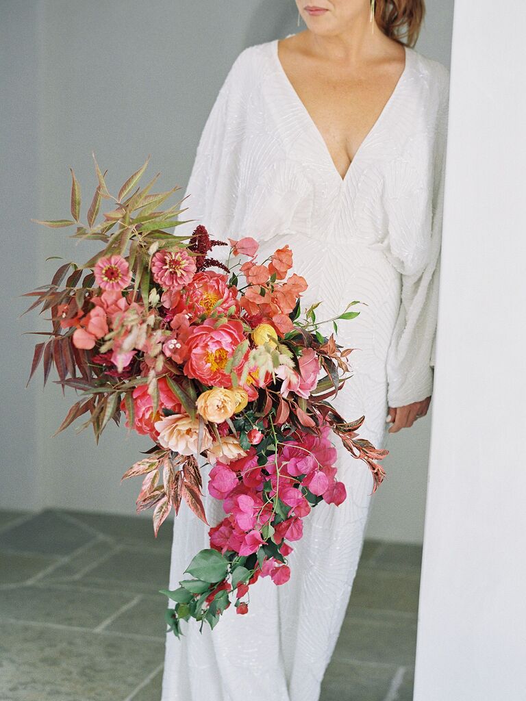 bride wearing white jumpsuit leans against wall holding colorful pink and orange cascading bouquet