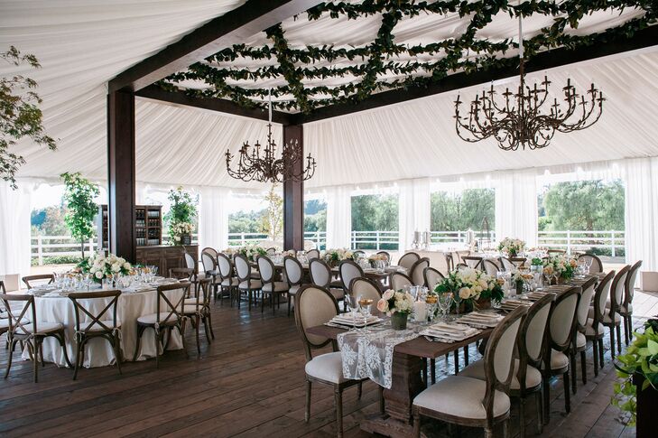 Under the elegant white tented reception space in Santa Barbara, California, dark wood beams connected to the ceiling that had elegant chandeliers dangling from above. Some dining tables had lace runners draped down the middle of bare wooden tops, while others had white tablecloths covering the entire surface.