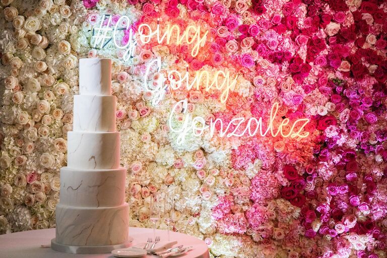 Five-tier wedding cake in front of flower wall with neon sign
