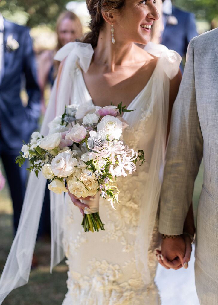 A bride with a fitted dress featuring tulle bows affixed to the shoulder straps carrying a light and elegant pastel bouquet filled with ivory, blush and blue flowers at her rustic-chic wedding.