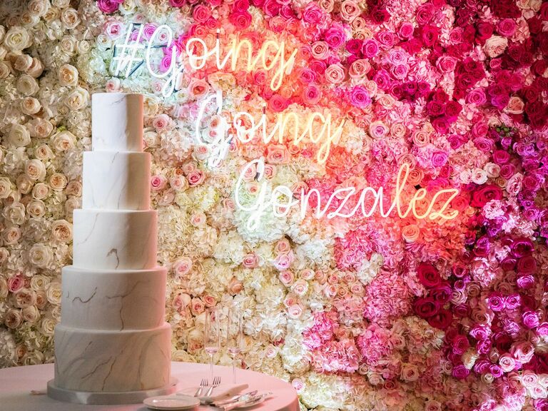 A marble white wedding cake in front of a neon wedding hashtag sign and flower wall. 
