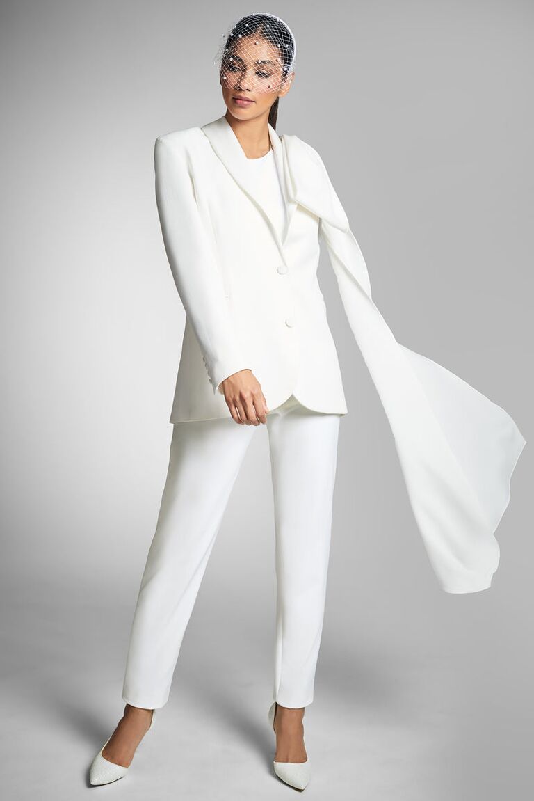 Model wears a white fitted pantsuit with an attached cape. 