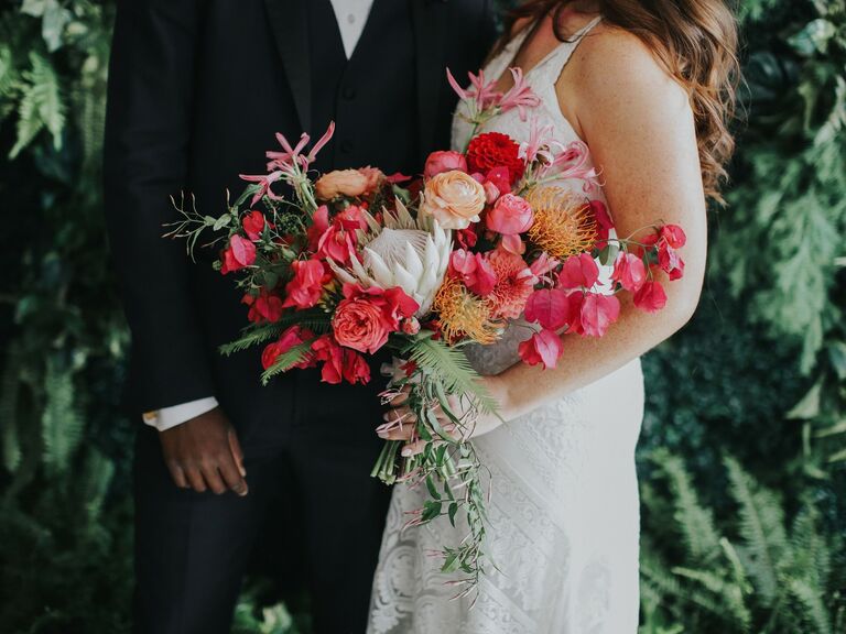 Bride and groom hold a vibrant pink bouquet featuring dahlias, roses, and greenery. 