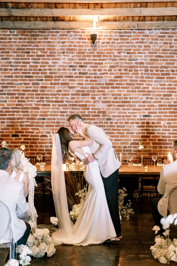 Groom Dipping Bride at Indoor Altar Against Brick Wall, Low Arrangements, First Kiss