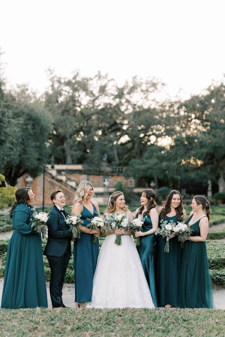 A bride in a whimsical, romantic A-line gown standing with her bridesmaids/wedding party, all in emerald green, in different styles of dresses, gowns and a suit with verdant bouquets featuring greenery and accented by white flowers at a sprawling villa in Miami, Florida.