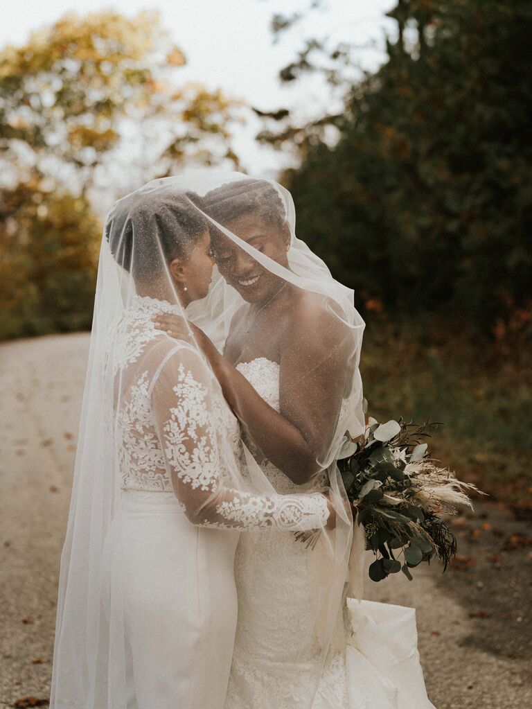 Brides holding each other under a large veil. 