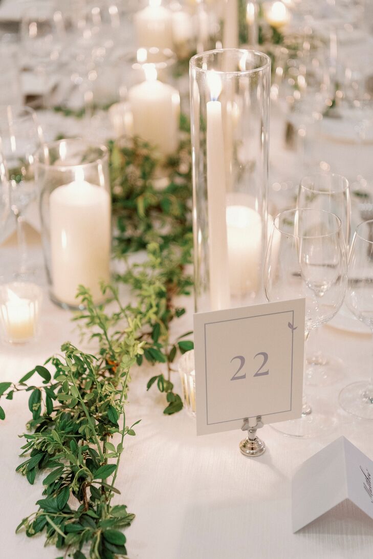A classic, simple and upscale white table setting with a simple table number, low and tall taper candles and a natural table runner with greenery and leaves winding down the table.