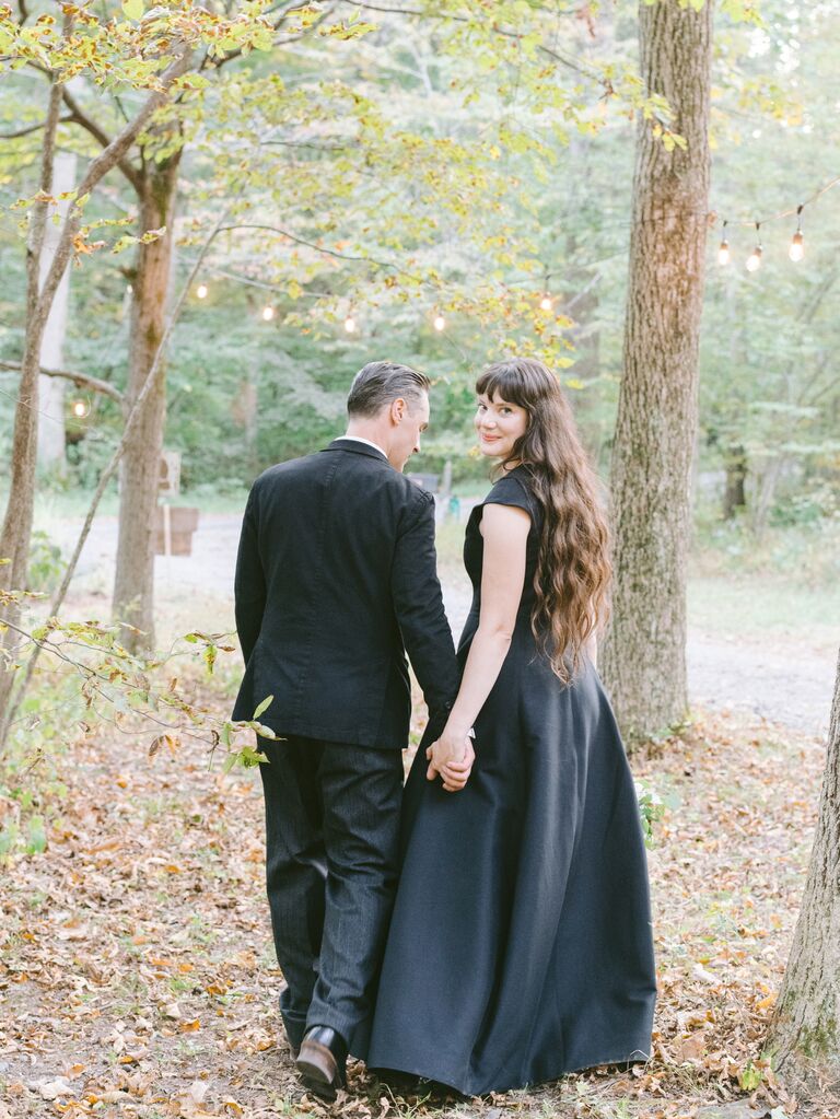 A woman turns back to look at the camera as she and her fiancé walk through the woods for their engagement photoshoot