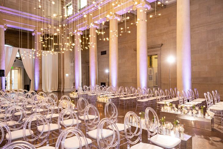 A ceremony space in the Baltimore Museum of Art featuring a hanging light installation, ghost chairs, elegant draping and minimalistic aisle decor with candles and low flower arrangements for a sophisticated, black-tie wedding.