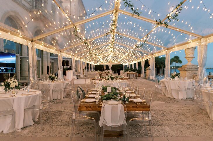 A romantic Jewish reception after sunset under a clear tent, string lights and suspended greenery featuring round tables with white linens and long tables with greenery runners with ghost chairs, gold accents and low centerpieces.