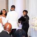 Bride in Off-the-Shoulder, Illusion Dress and Groom in White Tux at Altar With Officiant
