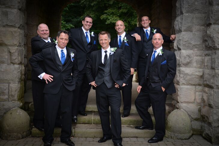The groom stood with his groomsmen, all dressed in black tuxedos. The groomsmen wore bright blue ties while the groom wore a black tie.