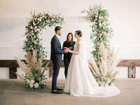 Bride and groom exchanging vows in front of lush floral arch and pampas grass