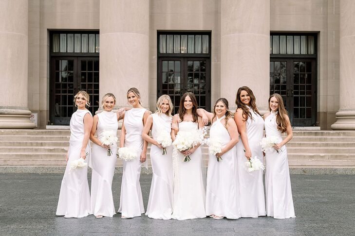 A bride in a fitted, modern gown and a cascading bouquet of all-white flowers standing on the steps of her venue with her bridesmaids in all-white gowns with white bouquets before her elegant, monochromatic wedding.