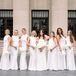 A bride in a fitted, modern gown and a cascading bouquet of all-white flowers standing on the steps of her venue with her bridesmaids in all-white gowns with white bouquets before her elegant, monochromatic wedding.