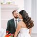 A bride with a bold bouquet of orange, red, yellow and lime green flowers and a glamorous, old-Hollywood down hairstyle of big waves with a groom in a deep olive suit in a museum hallway for their first look.