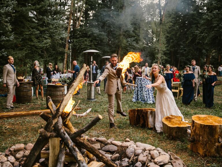 Newlyweds and the wedding party gather around a rustic bonfire in the woods. 