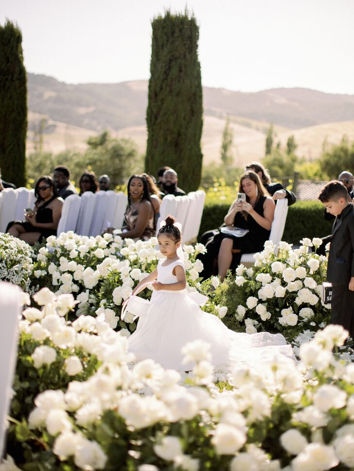 Flower Girl Walking Down Ceremony Aisle Surrounded by White Roses