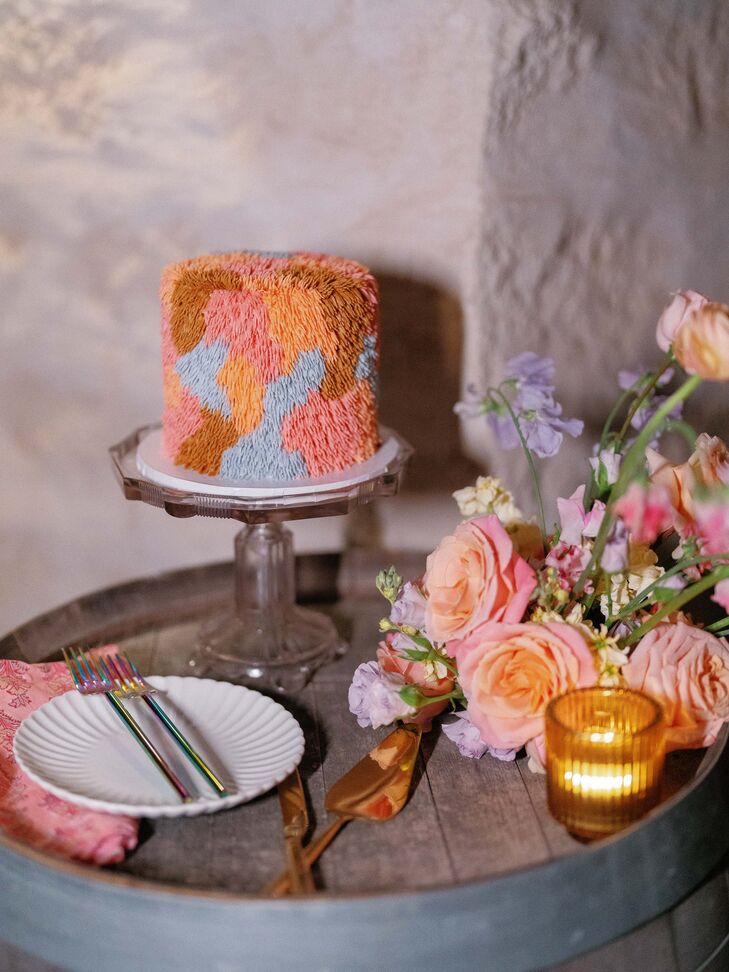 A single-tier, multi-colored, retro-inspired and "fuzzy" wedding cake featuring blue, orange, brown and pink colors on a dessert table with pastel flowers and low candles at a vintage-themed wedding.