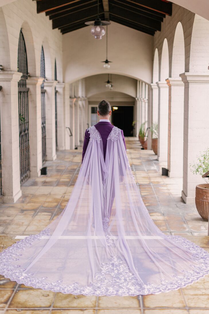 A groom in a unique, fashion-forward purple tuxedo showcasing his Cathedral-length tulle cape with intricate lace detailing on the hem, all in the style of a traditional veil.