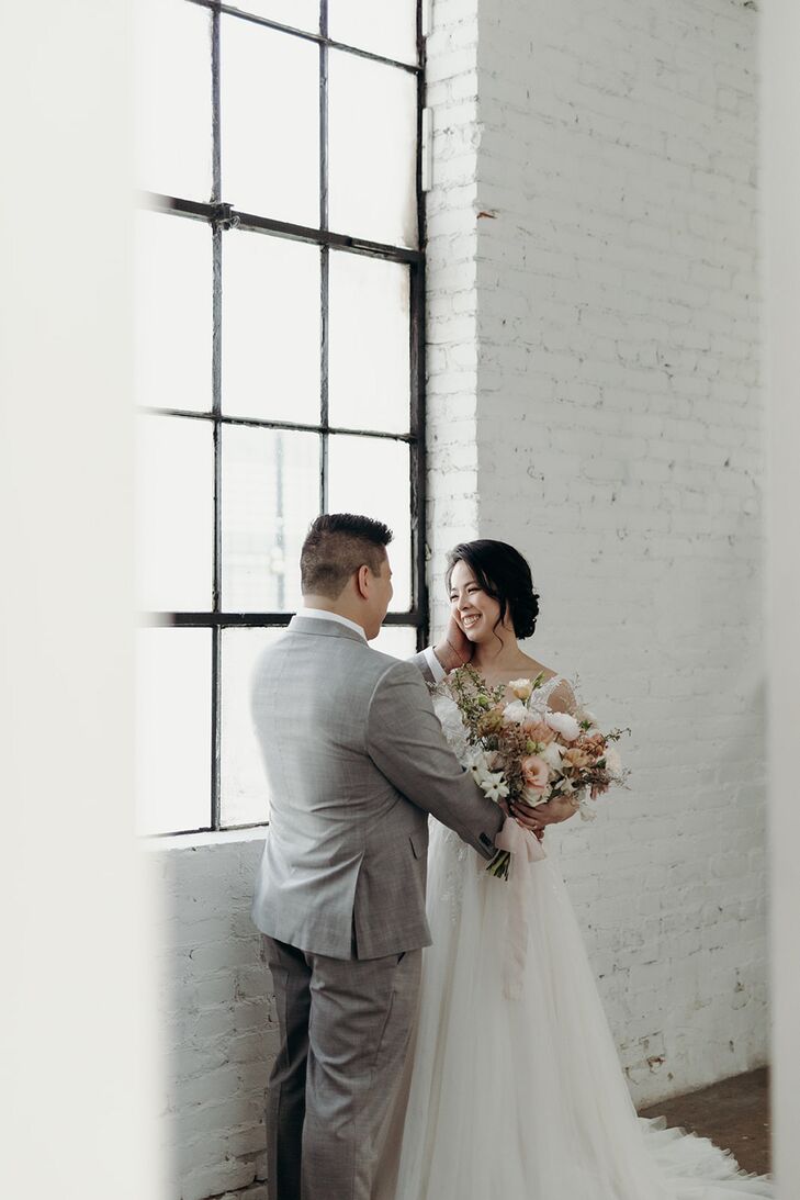 Groom in Gray Suit Embracing Bride in A-Line Gown at Window of Loft Venue, Pastels