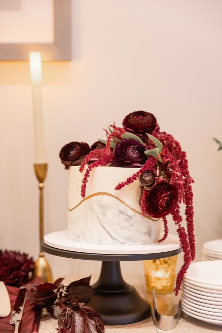 A single-tier white wedding cake with marble and gold designs topped with fresh marsala-wine-toned fresh flowers cascading down accented with greenery on a moody dessert table.