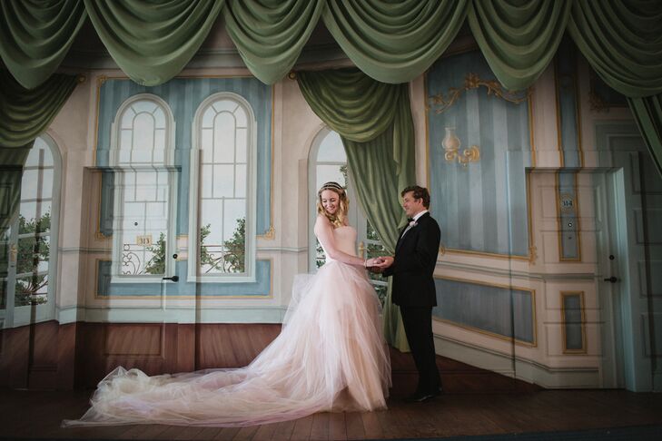 Bride in Blush Wedding Gown Holds Hands With Groom and Admires Train