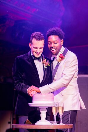 Grooms in Black-and-White Tuxedo Jackets Cutting Cake Under Purple Lighting
